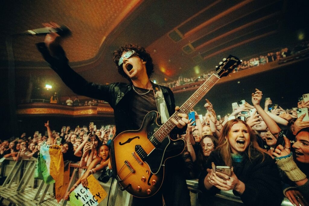Singer with an outstretched microphone and a guitar leaning toward a cheering crowd with phones raised